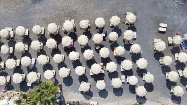 Aerial view of rows of white sun umbrellas and sunbeds on a pebble beach with people relaxing in Imperia, Liguria, Italy.