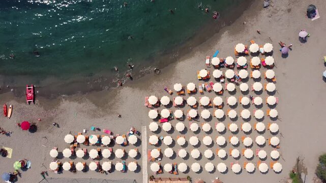 Aerial view of rows of white beach umbrellas on a sandy beach with people swimming in the turquoise sea in Imperia, Liguria, Italy.