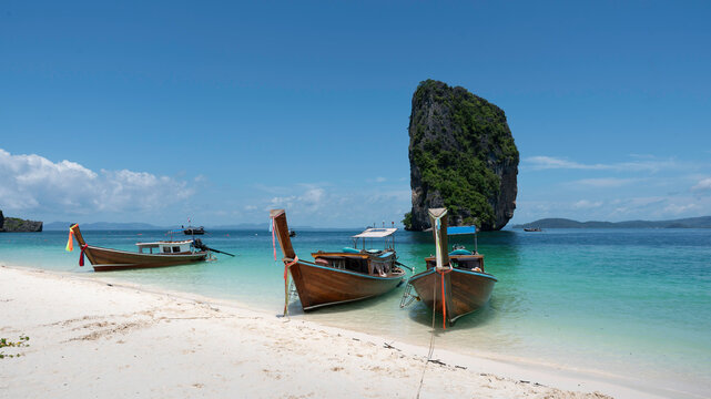 Beautiful tropical beach scene in Thailand featuring traditional wooden longtail boats resting on white sand beside crystal clear turquoise water. A dramatic limestone rock island rises from the sea u