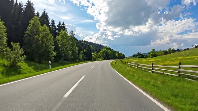 A long, empty road winds through a lush, green landscape under a bright, blue sky with clouds