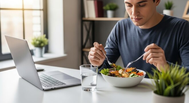 Man eating salad at desk with laptop. Mindful eating ritual during work break. Healthy lunch in office for professional wellbeing. Nutrition and healthy habit for student in workspace.
