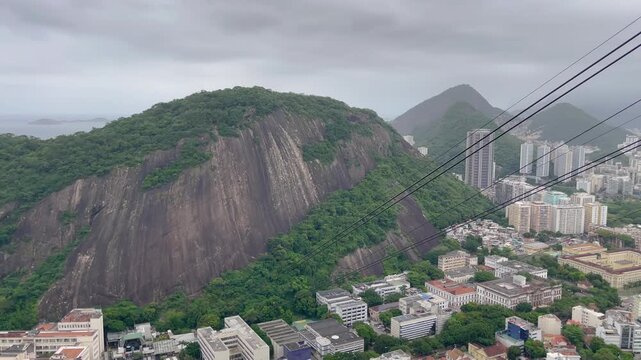 Slow panning cable car ride revealing Rio de Janeiro cityscape, dramatic granite peaks, and Guanabara Bay under heavy overcast skies.