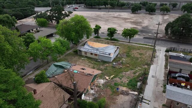 Damaged residential houses with destroyed roofs and blue tarps in Modesto, California, with green trees and nearby vacant lot under overcast sky in United States. Aerial drone top down