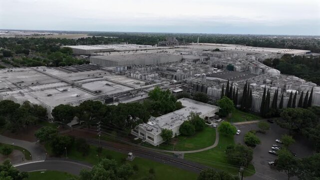 Industrial processing facility and warehouse buildings at Gallo Winery in Modesto, California, with lush green trees in foreground under overcast sky in United States. Aerial drone approaching shot.