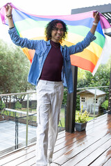 Fototapeta premium Non-binary person standing on wet deck holding rainbow pride flag, wearing denim overshirt