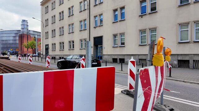 Urban construction site featuring tram rails stacked on the street, red and white safety barriers and bollards, with a light-colored residential building and industrial architecture in the background.