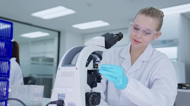 A scientist in a laboratory is focused on a microscope, looking closely at samples. The room is bright with modern equipment around her, and she wears a lab coat and gloves.