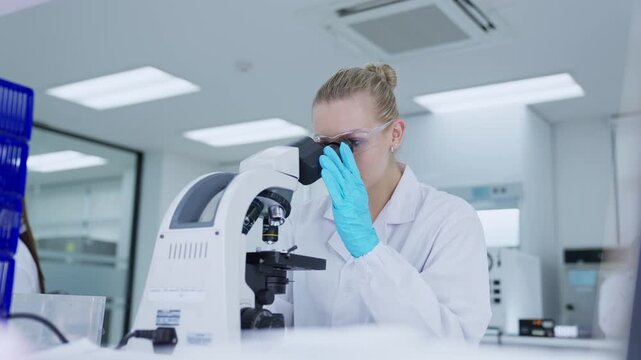 A scientist in a laboratory is focused on a microscope, looking closely at samples. The room is bright with modern equipment around her, and she wears a lab coat and gloves.