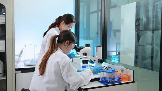 Two dedicated female scientists in lab coats working together, holding a clipboard and preparing liquid samples in test tubes during medical research