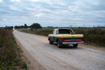 Vintage pickup truck driving on dirt road through rural countryside under cloudy sky © Matico