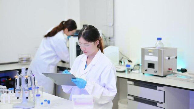Two dedicated female scientists in lab coats working together, holding a clipboard and preparing liquid samples in test tubes during medical research