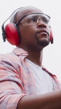 Vertical video: Reaching man in plaid shirt with red muffs and goggles raising arm outdoors to work
