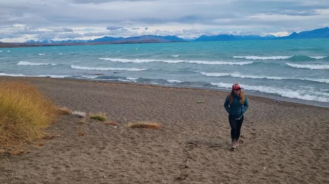 A female hiker returns along the sandy shore of Lago Argentino near El Calafate, Argentina, bundled in warm outdoor layers as strong Patagonian winds sweep across the remote lakeside landscape