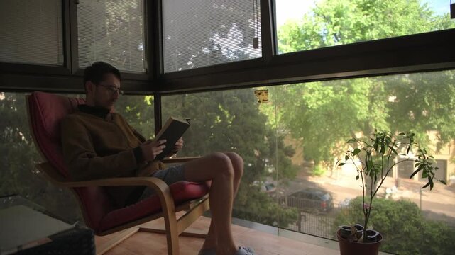 A man sits comfortably on a balcony chair, engrossed in a book. Sunlight filters through large windows, highlighting a lush garden outside. Ideal for lifestyle or relaxation themes.