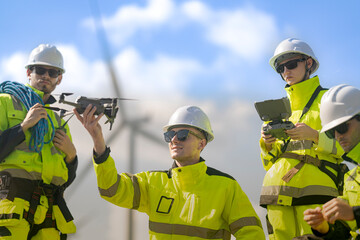 Fototapeta premium A group of engineers dressed in high-visibility uniforms and helmets conducts an inspection at a wind farm. Using technical devices, they monitor the wind turbines under a bright blue sky.