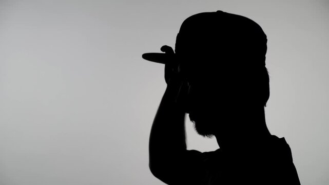 An anonymous silhouette of a man against a white wall, standing in profile, putting on and taking off a snapback cap