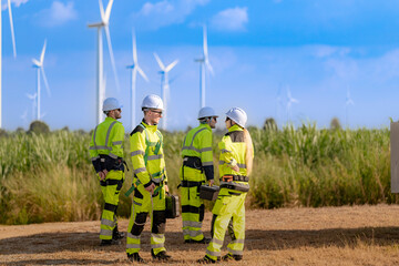 Naklejka premium A team of engineers in high visibility safety gear stand together at a wind farm, conducting inspections and discussing renewable energy projects. Wind turbines surround them under a clear sky.