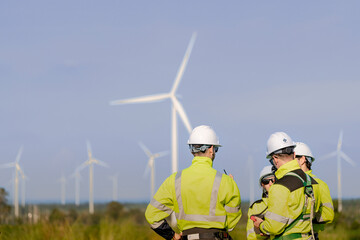 Fototapeta premium A team of engineers in high visibility safety gear stand together at a wind farm, conducting inspections and discussing renewable energy projects. Wind turbines surround them under a clear sky.