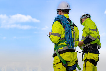 Fototapeta premium Two engineers in high visibility jackets and helmets are inspecting a wind farm. One is holding a mobile device, while the other is equipped with ropes for climbing.
