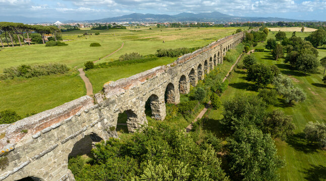 Aerial panoramic view of the Parco degli Acquedotti in Rome, Italy, featuring the long, ancient stone arches of a Roman aqueduct stretching across vibrant green fields toward the distant city skyline.