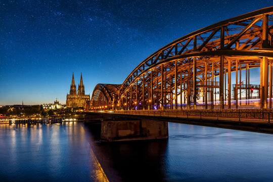 The illuminated cityscape of Cologne, Germany, with Hohenzollern Bridge and Cathedral during dusk with stars in the night sky