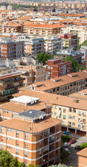 Obraz premium Aerial and vertical view of a dense urban residential area featuring numerous multi-story apartment buildings with terracotta roofs, solar panels, green trees, and parked cars in a sunny morning.