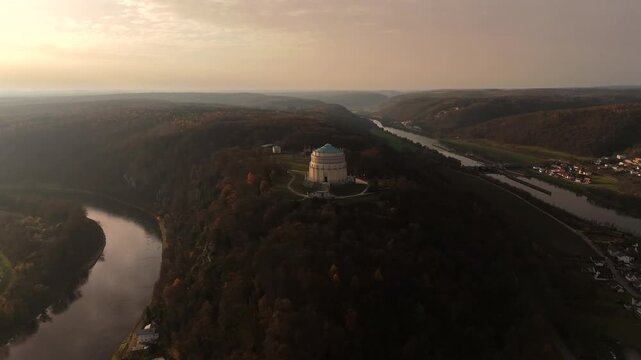 Panoramic drone view of Michelsberg hill with Hall of Liberation in Kelheim, Bavaria, Germany. Scene reveals Danube River, the Main-Danube Canal and dramatic fall foliage under warm sunset sunlight