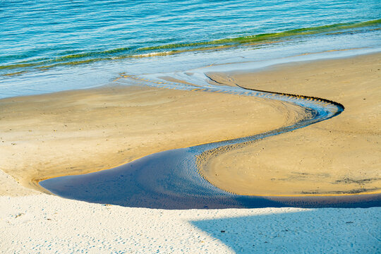 Keem Beach on Achill Island, County Mayo, Ireland, with calm blue water and a winding stream across the sand. This image shows the shoreline from land in warm light.