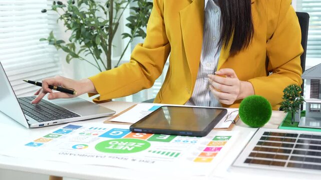 Businesswoman analyzing net zero and ESG sustainability reports with renewable energy model and eco-friendly office setup, focusing on green business strategy and carbon reduction.
