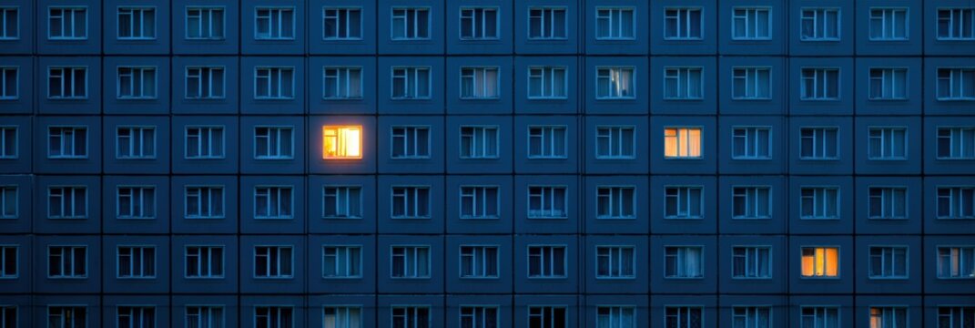 A warm light glows from one apartment window in a large dark building, standing out against the cold exterior in the night.