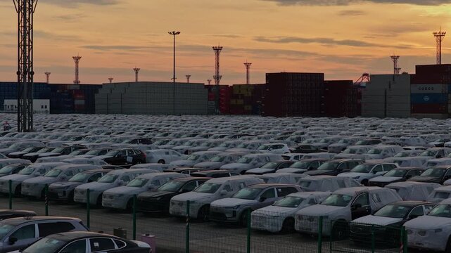 Rows of export cars in foreground with shipping containers and cranes at sunset