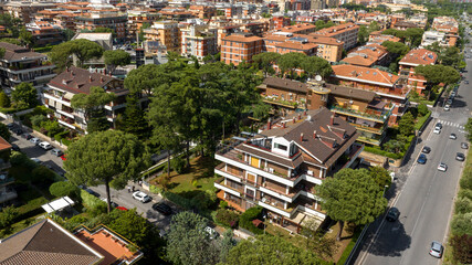 Fototapeta premium High-angle aerial view of a multi-story residential building featuring large balconies, nestled within a leafy neighborhood with tall pine trees, orange roofs, and a street with light car traffic.