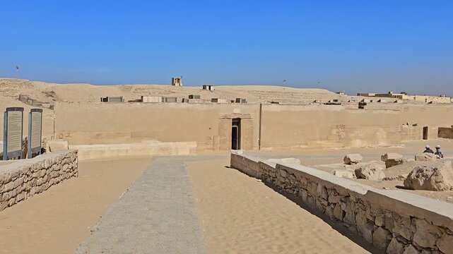 Exterior mud brick enclosure wall with carved stone doorway and relief figures at ancient Egyptian tomb of Kagemni at Saqqara necropolis, Egypt, sandy desert under blue sky.