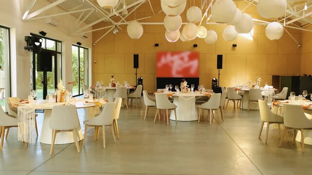 Wide shot of empty wedding banquet hall in boho style. Round white tables, grey chairs, paper lantern balls, audio speakers and big screen media display inside rustic venue. 4K slow motion.
