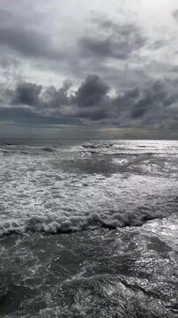 A turbulent ocean scene with dark clouds overhead. Waves crash against the shore, creating a foamy surface. The sky is overcast, reflecting a moody atmosphere.