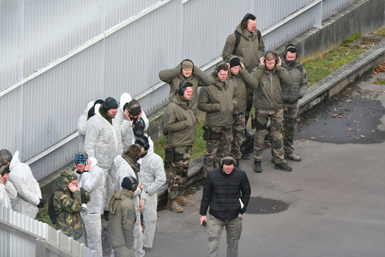 Group of soldiers and trainees in camouflage and white coveralls stand outdoors near a fence, many wearing hearing protection and tactical gear during a training exercise