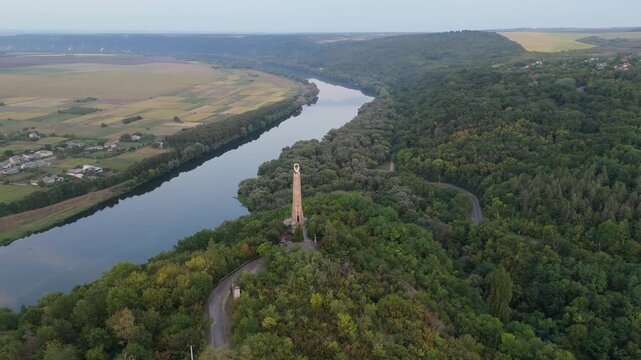 Panoramic aerial view of the Thanksgiving Candle monument in Soroca, Moldova. Drone flight over lush green hills and the Dniester River valley at sunset, featuring the iconic stone landmark.