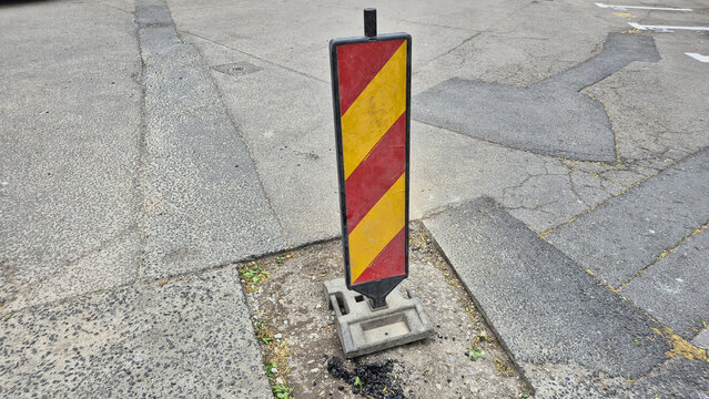 A vertical, rectangular traffic delineator with red and yellow diagonal stripes, standing on a grey weighted base in the middle of a paved asphalt surface.
