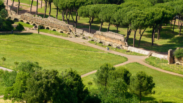 Aerial view of the Parco degli Acquedotti in Rome, Italy, showcasing ancient ruins alongside lush greenery. Stone pine trees and walking paths surround the weathered remains of a historical Roman city