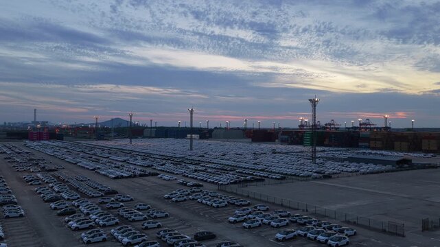 Wide aerial view of automobile storage yard and city lights near port after sunset