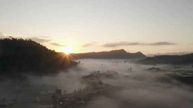 Aerial view of the sun rising over a thick layer of fog covering a rural valley and silhouetted hills in Batur, Central Java, Indonesia.