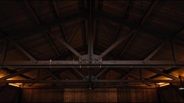 Interior View of Rustic Wooden Ceiling Structure with Heavy Timber Beams and Exposed Trusses in Dark Barn or Warehouse Setting with Warm Ambient Lighting and Industrial Architecture Detail