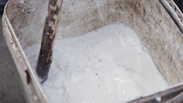 A wooden stick mixes thick white lime whitewash in a plastic bucket for seasonal tree bark protection to prevent sunscald damage and stop harmful insect infestations in the garden
