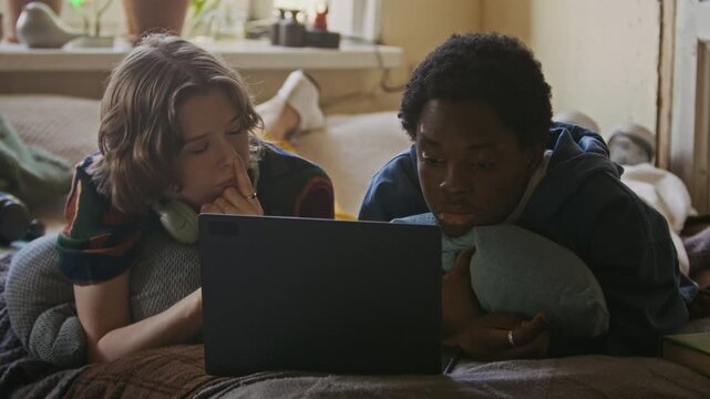 Medium close up shot of couple of diverse gen Z freelancers working on laptop while lying on bed in shared apartment