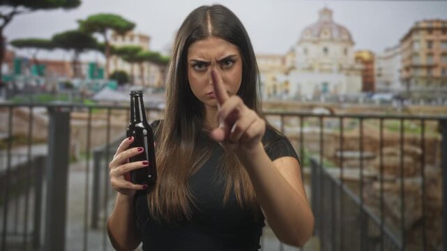 Woman holding beer bottle and pointing finger at camera by historic building plaza, stern expression and pointing gesture; anger defiance confrontation.