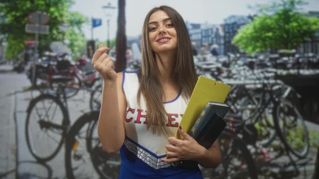 Woman cheerleader holding yellow clipboard and stacked books makes finger heart gesture facing camera on street with parked bicycles and blurred buildings; playful charm.