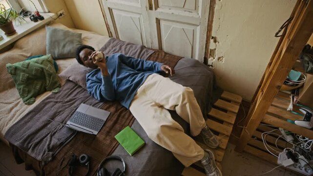 View from above of gen Z African American man lying in bed, throwing baseball and working on laptop from apartment