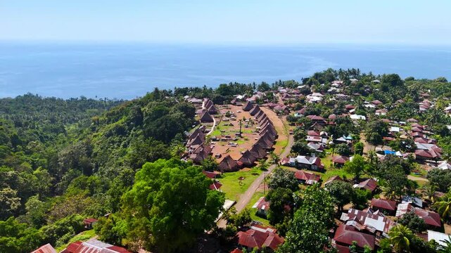 Aerial view of Kampung Adat Nggela traditional village with unique thatched roofs on a lush green hill overlooking the blue sea in Wolojita, East Nusa Tenggara, Indonesia.