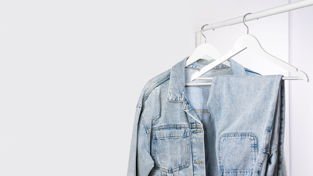 Light blue denim jacket, jeans on white hangers on rack, gray background. Concept of minimalist wardrobe, simple fashion aesthetic, capsule clothing and conscious consumption lifestyle. Copy space
