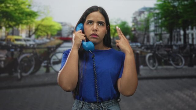 Young woman holding blue handset to ear, hand raised and rolling eyes on a street with canal and bicycles; frustration miscommunication.